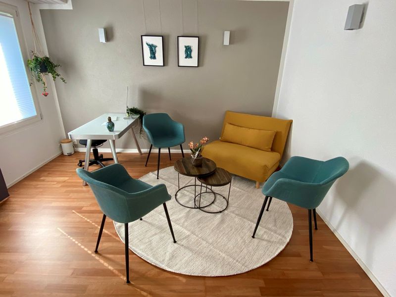 Heller Beratungsraum: drei petrolblaue Sessel und ein senfgelbes Sofa um zwei kleine Holztische auf einem runden Teppich, dahinter Schreibtisch und Fenster